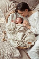 Mother next to a newborn in a wicker basket under the all-season Meri merino blanket in warm beige