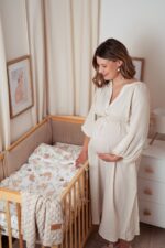 Pregnant mother by a cot with Forest Friends bedding, Fema bamboo blanket in warm beige and Velvet bumper in cappuccino