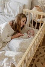 Mama przy łóżeczku z niemowlęciem przykrytym kremową pościelą Herbs w drobne gałązki