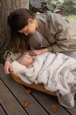 Mama pochyla się nad niemowlęciem w koszu przykrytym kocykiem bawełnianym Birdsong