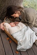 Mother leaning over a baby in a basket covered with the Happy Goose Olive blanket