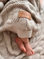 Zbliżenie na stópki niemowlęcia i ażurowy splot beżowego kocyka bambusowego Ivi z metką