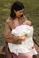 Mama w parku trzyma niemowlę owinięte kremowym kocykiem bambusowym Hana