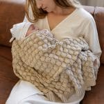 Mother on a sofa holding a newborn in a hat wrapped in the Fema bamboo blanket in beige
