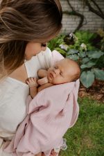 Mama w ogrodzie tuli noworodka otulonego różową pieluszką muślinową