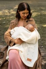 Mama siedzi na trawie i tuli niemowlę w kremowym kocyku merino Meri