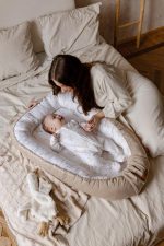 Mother holding the hand of a baby lying in the beige Happy Goose baby nest on a bed