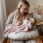 Mama z niemowlęciem na poduszce do karmienia, dziecko przykryte różową pieluszką muślinową