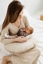 Mama siedzi na łóżku z noworodkiem na poduszce do karmienia Herbs