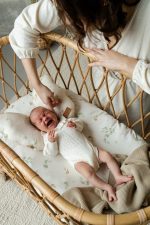 Mother adjusting the pillow of a crying newborn on a Eucalyptus sheet in a rattan crib