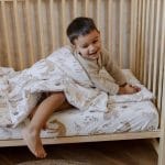 Smiling boy climbing out from under the Forest Friends duvet in a wooden cot
