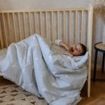 Laughing boy tucked into the Happy Goose Olive duvet in a wooden cot