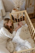 Mama otula niemowlę beżową kołdrą Birdsong w drewnianym łóżeczku