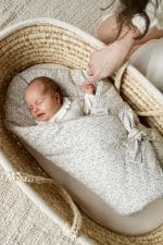 Sleeping newborn in the Herbs swaddle wrap in a wicker basket as mother adjusts the bow