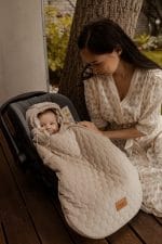 Mama pochyla się nad niemowlęciem w foteliku otulonym beżowym śpiworkiem Herbs