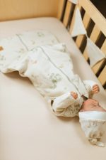 Noworodek w śpiworku Eucalyptus w kolorze ecru leży w drewnianym łóżeczku