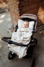 Smiling baby in a stroller by a brick wall under the Meri merino blanket in warm beige
