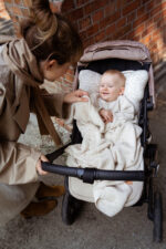 Smiling baby in a stroller tucked in the Meri merino blanket in warm beige, mother leaning in