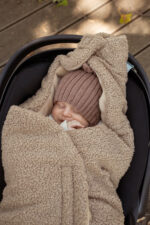 Close-up of a baby in a pink knitted hat under the brown Teddy car seat swaddle with the hood fastened